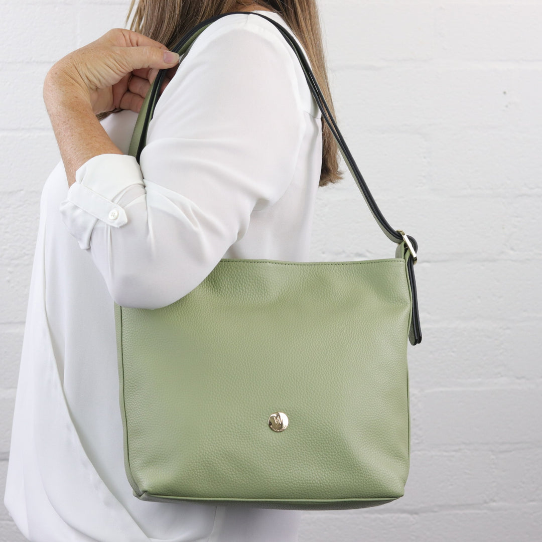 woman wearing sage green coloured leather bag on shoulder with wide short strap and gold buckle and badge logo#colour_sage