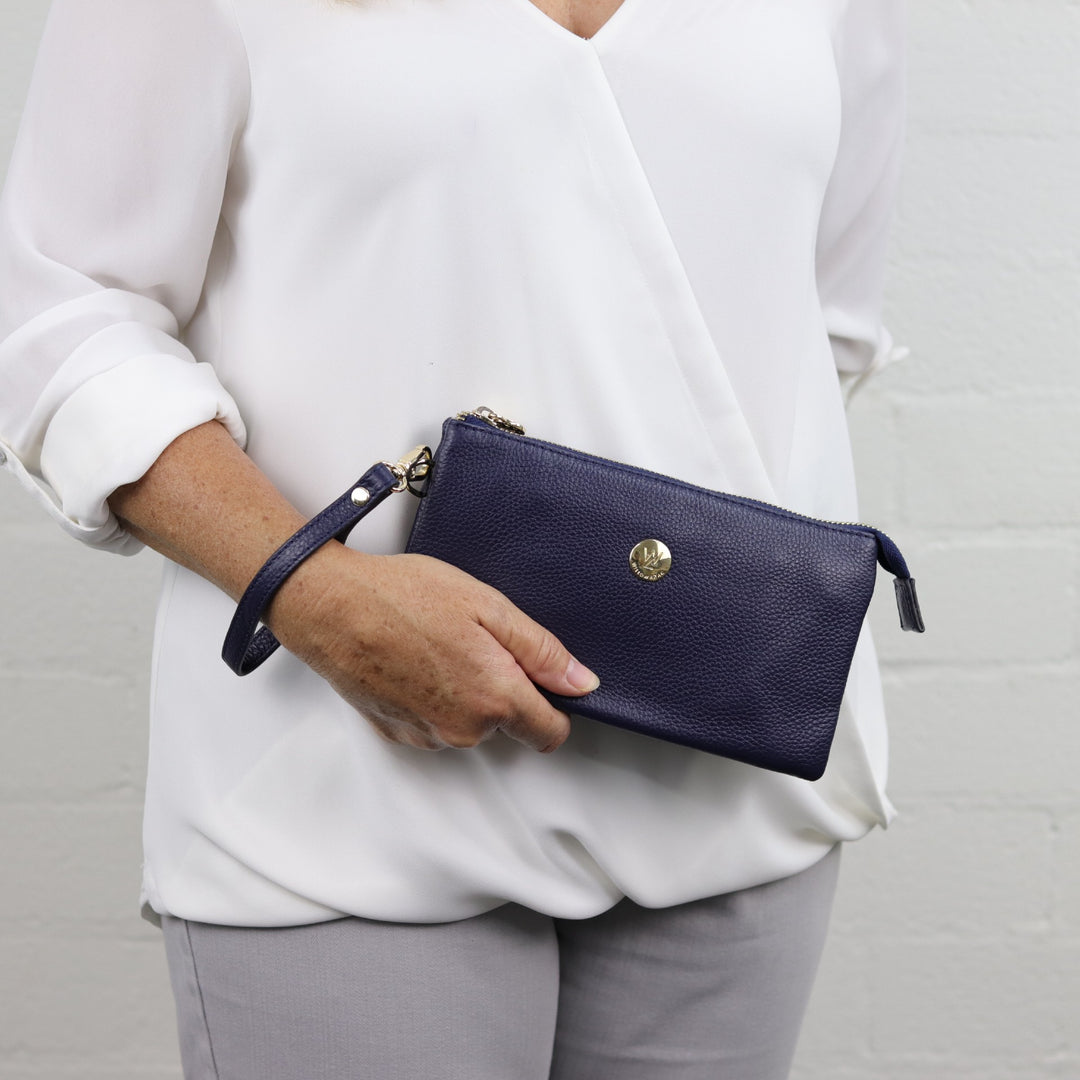 woman holding navy leather clutch with gold hardware and attached wrist strap#colour_navy