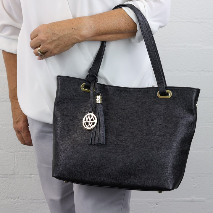 woman wearing black pebbled leather tote handbag on arm with double handles and tassel with gold logo charm #colour_black