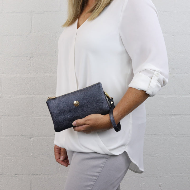 woman model holding shiny metallic navy leathe clutch with gold button logo and zip closure#colour_raven